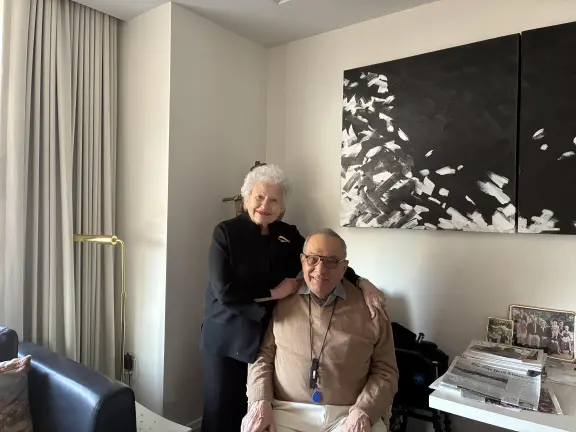 Rosalyn and Irwin Engelman in their living room. She stands behind him with hands on his shoulders; both smile warmly.. 